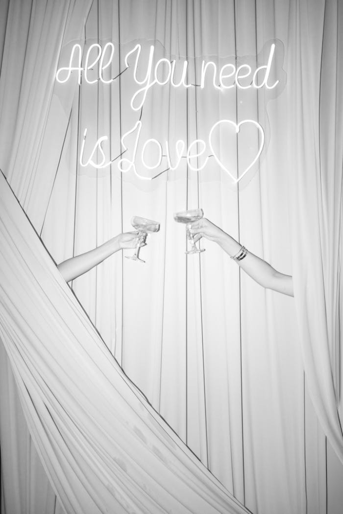 Two people toasting with champagne under 'All You need is love' neon sign.