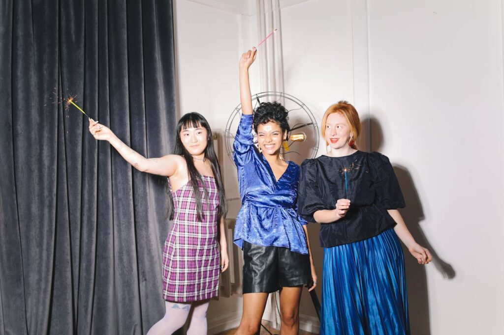 Three women joyfully celebrating indoors with sparklers, showcasing diverse fashion styles.